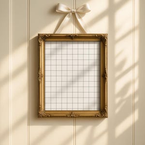 Op de afbeelding: Een goudkleurige, sierlijke fotolijst met een rasterpatroon, hangend aan een cr&egrave;mekleurig lint dat in een strik is geknoopt. De lijst is rechthoekig en heeft decoratieve details. De achtergrond is een lichtbeige muur.