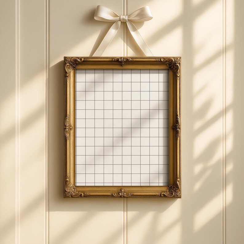 Op de afbeelding: Een goudkleurige, sierlijke fotolijst met een rasterpatroon, hangend aan een cr&egrave;mekleurig lint dat in een strik is geknoopt. De lijst is rechthoekig en heeft decoratieve details. De achtergrond is een lichtbeige muur.