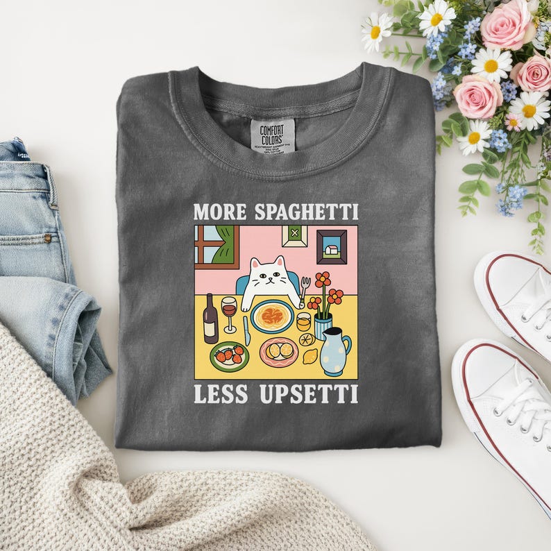 Puede incluir: Camiseta gris oscuro con el texto "MORE SPAGHETTI LESS UPSETTI" y un gr&aacute;fico de un gato en una mesa puesta. El gato est&aacute; sentado en una mesa con espaguetis, vino y flores. La camiseta es de Comfort Colors.