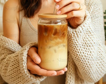 Op de afbeelding: Een persoon houdt een glazen pot met een rietje vast, gevuld met ijskoffie. De pot heeft een houten deksel en de koffie is gelaagd met een lichtbruine bovenkant en een donkerbruine onderkant.