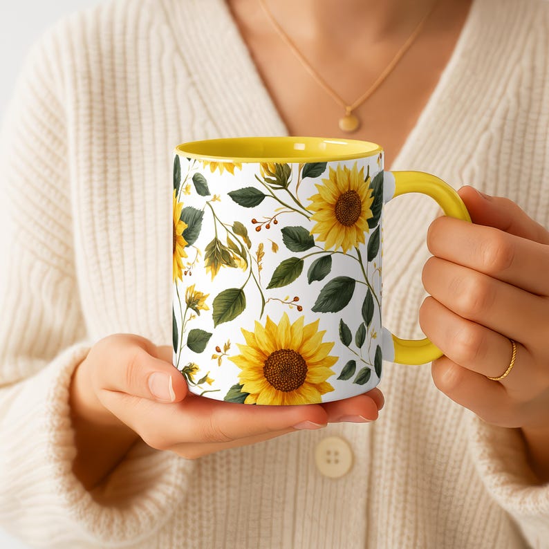 May include: A white ceramic mug with a yellow interior and handle, decorated with a sunflower pattern. The design features large yellow sunflowers, green leaves, and floral accents. The mug is held in a person's hands.