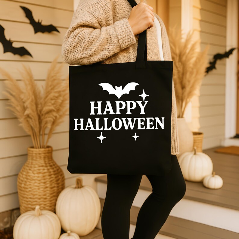 Op de afbeelding: Een zwarte draagtas met de witte tekst "HAPPY HALLOWEEN", samen met vleermuis- en sterrenafbeeldingen. De tas wordt door een persoon vastgehouden. De achtergrond omvat pompoenen, decoratief gras en vleermuisuitsparingen, waardoor een feestelijke Halloween-sc&egrave;ne ontstaat.