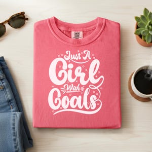May include: A folded coral-colored t-shirt with the text "Just A Girl With Goals" in white script. The shirt is on a light wood surface, with sunglasses, jeans, a succulent, and a cup of coffee nearby.