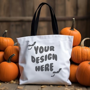 Może przedstawiać: Biała torba typu tote z czarnymi uchwytami, z napisem "YOUR DESIGN HERE" i sylwetkami nietoperzy. Torba otoczona jest kilkoma pomarańczowymi dyniami, tworząc motyw jesienny lub Halloween.