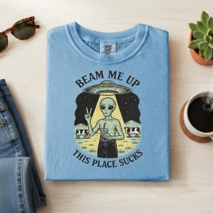 May include: Light blue t-shirt with a graphic of an alien holding a drink, giving a peace sign, and standing under a UFO. The text reads "Beam Me Up, This Place Sucks". The shirt is folded.