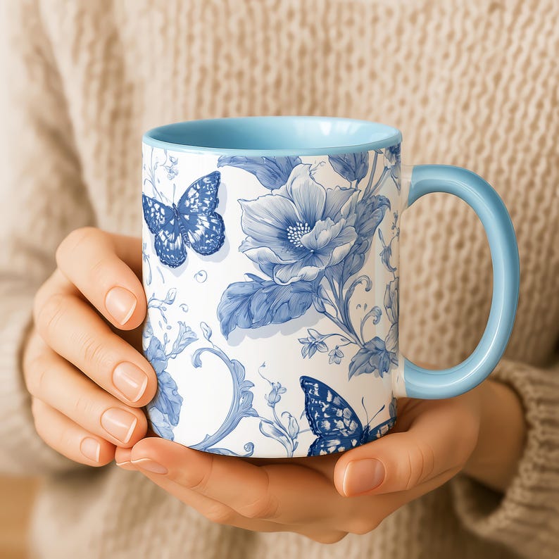 May include: White ceramic mug with a blue floral and butterfly pattern. The mug has a light blue interior and handle. The design features detailed blue flowers and butterflies on a white background, creating a classic, elegant appearance.