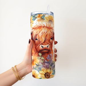 May include: A stainless steel tumbler with a colorful design featuring a Highland cow with sunflowers and other flowers. The tumbler has a straw and is being held by a hand.
