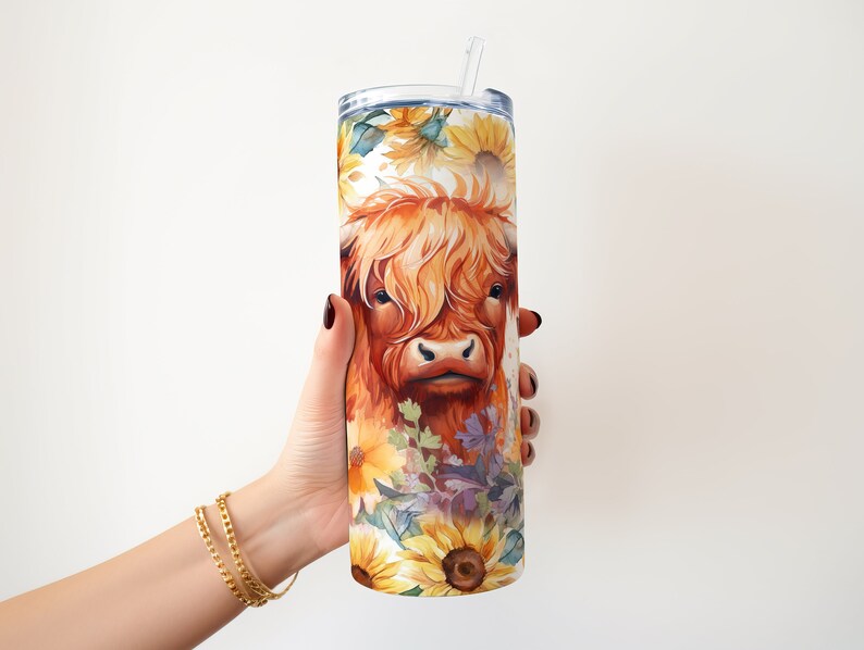 May include: A stainless steel tumbler with a colorful design featuring a Highland cow with sunflowers and other flowers. The tumbler has a straw and is being held by a hand.