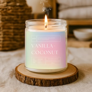 Op de afbeelding: Een brandende vanille- en kokosgeurkaars in een heldere glazen pot, op een houten onderzetter. Het kaarslabel bevat de tekst "Vanilla + Coconut". De kaars is 9 cm hoog.