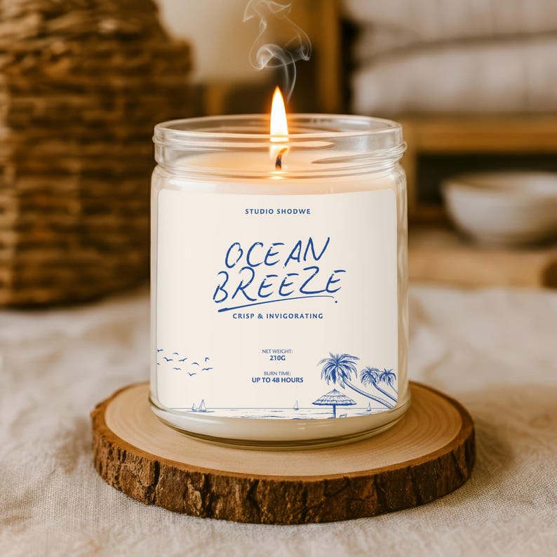 Op de afbeelding: Een brandende glazen potkaars met de tekst "Ocean Breeze" en een strandillustratie. De kaars staat op een houten onderzetter. Nettogewicht 210g, brandtijd tot 48 uur.