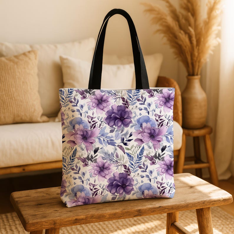 Puede incluir: Una bolsa de tela blanca con un estampado floral con flores de acuarela moradas y azules. La bolsa tiene asas negras.