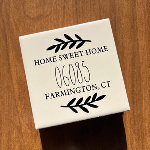 Op de afbeelding: Witte keramische tegel met zwarte tekst "Home Sweet Home 06085 Farmington, CT" met een zwart bloemmotief boven en onder de tekst.