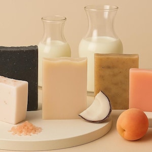 Peut inclure: Assortiment de savons artisanaux aux couleurs et textures variées, dont un marbré gris, un noir uni et une barre rose clair. Les savons sont présentés avec des abricots, une tranche de noix de coco et de petits éléments décoratifs.