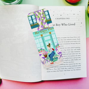 May include: A decorative bookmark depicting a Parisian cafe, with the text "Au Vieux Paris". The illustration shows a woman on a bicycle, purple flowers, and a teal facade. The bookmark is placed in an open book.