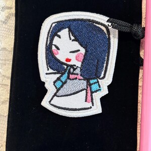 May include: Embroidered patch featuring a cartoon illustration of a woman with dark blue hair, pink cheeks, and red lips. The patch is outlined in white and is on a black velvet pouch. The woman is wearing a light blue and pink outfit.