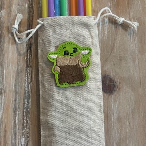 May include: A beige drawstring bag with a green and brown embroidered character patch. The bag contains several colorful reusable straws in yellow, purple, blue, green, and orange. The bag is made of a textured fabric.