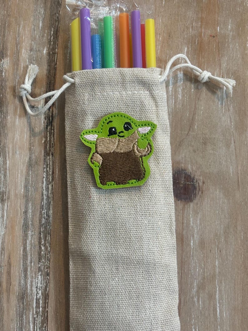 May include: A beige drawstring bag with a green and brown embroidered character patch. The bag contains several colorful reusable straws in yellow, purple, blue, green, and orange. The bag is made of a textured fabric.
