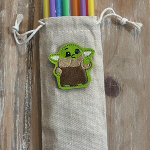 May include: A beige drawstring bag with a green and brown embroidered character patch. The bag contains several colorful reusable straws in yellow, purple, blue, green, and orange. The bag is made of a textured fabric.