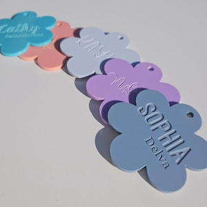May include: Five flower-shaped tags with personalized names. The tags are in various pastel colors, including blue, pink, purple, and light blue. The names on the tags are "Cally", "May", "Nola", and "Sophia Delva".