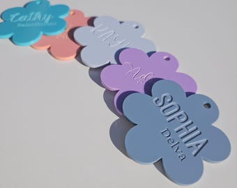 Puede incluir: Cinco etiquetas en forma de flor con nombres personalizados. Las etiquetas son de varios colores pastel, incluyendo azul, rosa, morado y azul claro. Los nombres en las etiquetas son "Cally", "May", "Nola" y "Sophia Delva".