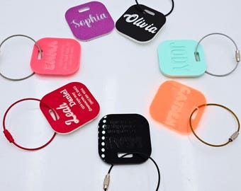 May include: Six colourful square luggage tags with names printed on them. Each tag has a metal ring attached to it. The tags are pink, red, purple, black, orange, and turquoise.