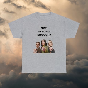 May include: Grey t-shirt featuring a graphic of three people pointing at the viewer. The text "NOT STRONG ENOUGH?" is printed above the graphic.