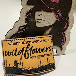 May include: Wooden sign with the text "where others see weeds wildflowers see opportunity" and a silhouette of a person in a field. A separate wooden cutout features a woman wearing a hat with "MAD ACRE FARMS" printed on it.