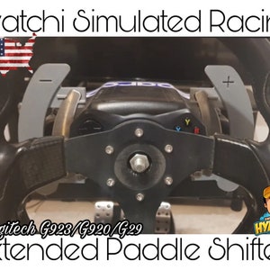 May include: A close-up of a black Logitech G923/G920/G29 racing wheel with extended paddle shifters. The paddles are grey and have plus and minus signs on them. The wheel has a black leather grip and a silver centre hub.