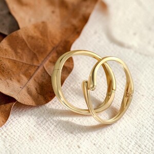 May include: Two gold-coloured hoop earrings interlocked, set against a textured white surface. The earrings have a smooth, polished finish and a unique design. A dried brown leaf is visible in the background.