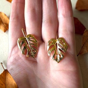 May include: A pair of gold-coloured earrings shaped like Monstera leaves. The earrings have a textured surface and a hook closure. The earrings are displayed on a hand, with dried leaves in the background.