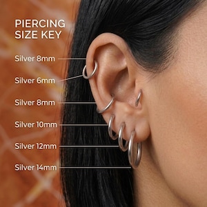 May include: A silver hoop earring size guide featuring text PIERCING SIZE KEY and labels for silver hoops ranging from 6mm to 14mm. The image displays multiple silver hoop earrings of varying diameters positioned along the ear cartilage and lobe.