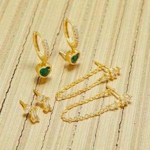 Puede incluir: Una colección de pendientes dorados. Dos pares de pendientes presentan una piedra verde en forma de corazón y una pequeña cadena. El otro par de pendientes son pequeños tachuelas con una piedra clara rectangular.