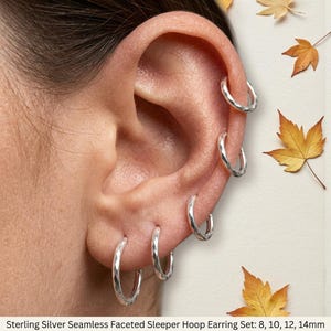 May include: A set of five sterling silver seamless faceted sleeper hoop earrings in sizes 8mm, 10mm, 12mm, and 14mm. The hoops feature a textured, reflective surface and are arranged along the outer edge of an ear, showcasing their circular design.
