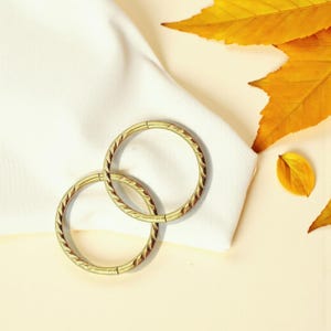 Può includere: Due anelli circolari in metallo color oro con un design intrecciato strutturato. Gli anelli sono collegati e poggiano su un tessuto bianco, con foglie autunnali gialle sullo sfondo. Probabilmente si tratta di gioielli o forniture artigianali.