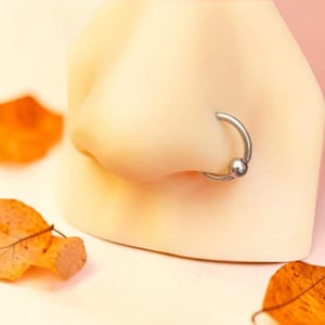 May include: A silver nose ring with a small ball closure is shown on a mannequin nose. The nose ring is a hoop style, and the ball is located at the bottom of the hoop. The background is a soft, neutral colour.