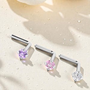 以下が含まれることがあります： 3つのシルバーL字型ノーズスタッド、宝石アクセント付き。宝石は紫、ピンク、クリアです。スタッドは明るい色の表面に表示されています。