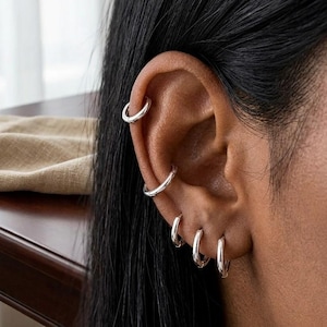 May include: A set of five polished silver hoop earrings worn in a single ear. The collection includes one hoop in the upper cartilage, one in the mid-cartilage, and three smaller hoops arranged along the lower earlobe in a vertical row.