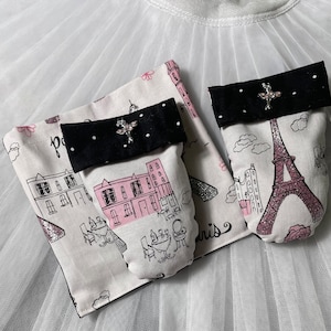 Puede incluir: Un par de guantes de horno de tela blanca con un diseño de temática parisina en blanco y negro. Un guante presenta una Torre Eiffel rosa y el otro un edificio rosa con un toldo blanco y negro. Ambos guantes tienen un ribete blanco y negro.