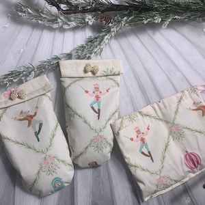 May include: Three small white fabric gift bags with a Nutcracker theme. The bags have a white background with a green and pink floral pattern and a Nutcracker illustration. The bags are on a white tulle fabric.
