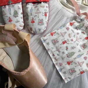 May include: A pair of ballet pointe shoes with satin uppers and ribbons, next to two fabric toe pads and a drawstring bag. The fabric features a holiday print with ballerinas, nutcrackers, and Christmas trees. The toe pads have red tops with a Christmas tree charm.