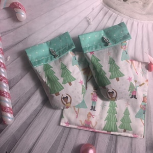 May include: Two small green and white fabric gift bags with a Christmas tree pattern. The bags have a silver charm attached to the top. The bags are sitting on a white tulle fabric.
