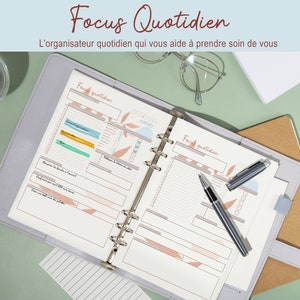 May include: A gray and white daily planner with a ring binder. The planner has the words "Focus Quotidien" and "L'organisateur quotidien qui vous aide &agrave; prendre soin de vous" printed on the cover. The planner is open to a page with the words "Face quotidien" printed at the top.