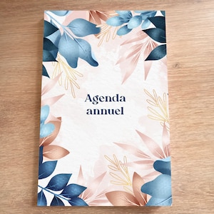 Peut inclure: Une couverture de carnet d'agenda blanche avec un motif floral. Le motif présente des feuilles bleues et roses avec des accents dorés. Le texte "Agenda annuel" est imprimé en bleu sur la couverture.