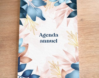 May include: A white agenda book cover with a floral design. The design features blue and pink leaves with gold accents. The text "Agenda annuel" is printed in blue on the cover.