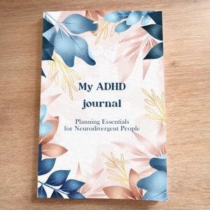 Peut inclure: Une couverture de journal blanche avec le texte "My ADHD journal" et "Planning Essentials for Neurodivergent People". La couverture présente un motif floral avec des feuilles bleues, roses et dorées.