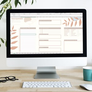 May include: A computer screen displaying a digital planner with the title "Focus Quotidien". The planner has sections for daily tasks, goals, and notes. The screen is surrounded by a white background and a wooden desk with a keyboard, mouse, and a blue mug.