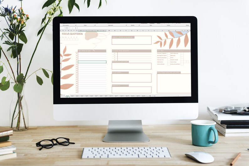 May include: A computer screen displaying a digital planner with the title "Focus Quotidien". The planner has sections for daily tasks, goals, and notes. The screen is surrounded by a white background and a wooden desk with a keyboard, mouse, and a blue mug.