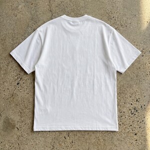 Puede incluir: Una camiseta blanca lisa de manga corta. La camiseta est&aacute; hecha de una tela suave y opaca y tiene cuello redondo. La parte trasera de la camiseta es visible, mostrando el dise&ntilde;o y la construcci&oacute;n simples.