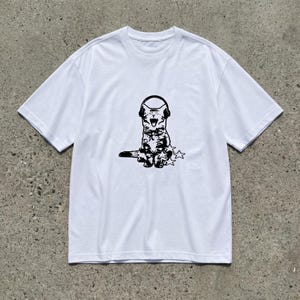 Puede incluir: Camiseta blanca con un dibujo negro de un gato con auriculares y tres estrellas. El gato tiene la boca abierta. La camiseta tiene mangas cortas y cuello redondo. La prenda es de material suave.