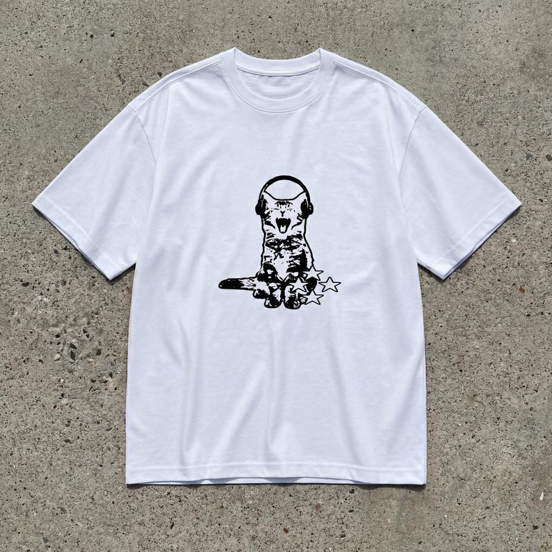 Puede incluir: Camiseta blanca con un dibujo negro de un gato con auriculares y tres estrellas. El gato tiene la boca abierta. La camiseta tiene mangas cortas y cuello redondo. La prenda es de material suave.